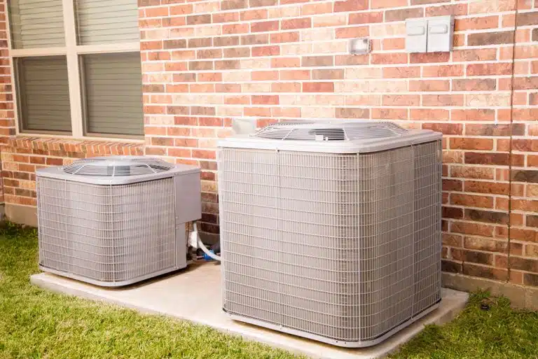 Two HVAC units, one larger and one smaller, installed on a concrete slab outside a brick home.