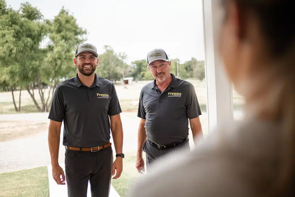 Two Frymire technicians smiling while greeting a homeowner at the front door. View from over the homeowner's shoulder.