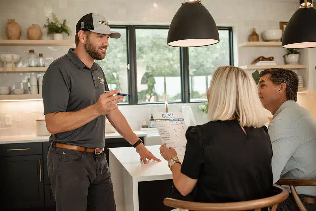 A technician standing in a homeowner's kitchen, speaking to the homeowners who are sitting at their kitchen island. Kitchen is updated style.