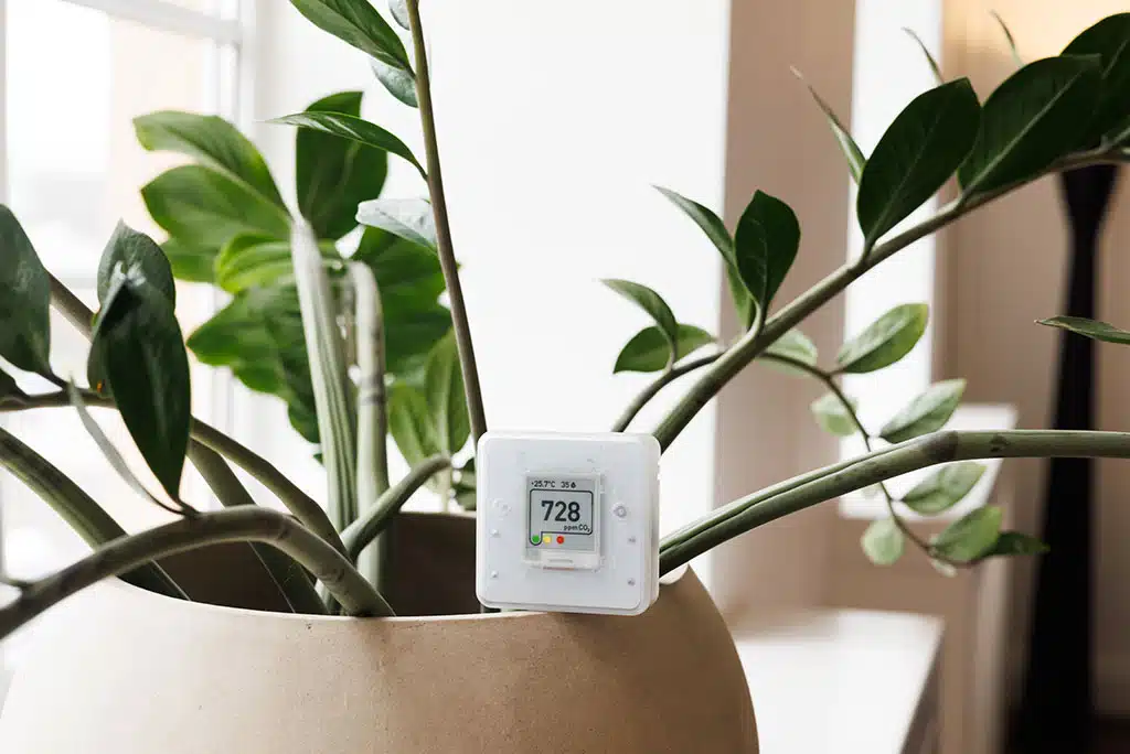 Close-up of an air quality index reading device sitting on the edge of a potted plant in a home.