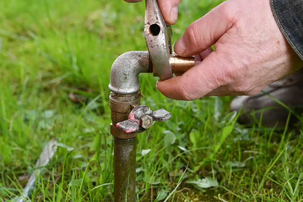 Close-up of hands holding and applying wrench to an outdoor faucet in green grass.