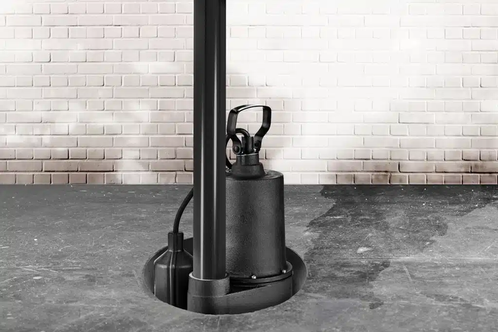A black sump pump installed in a basement floor with white brick wall in background.