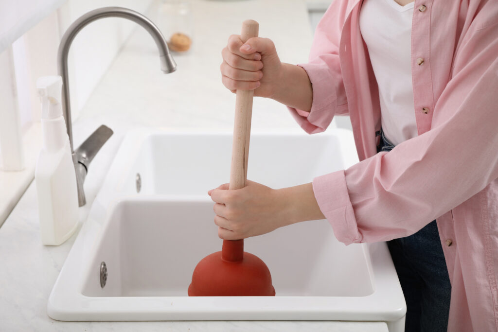 Woman using plunger to unclog sink drain in the kitchen. White porcelain double sink with brushed nickel faucet.